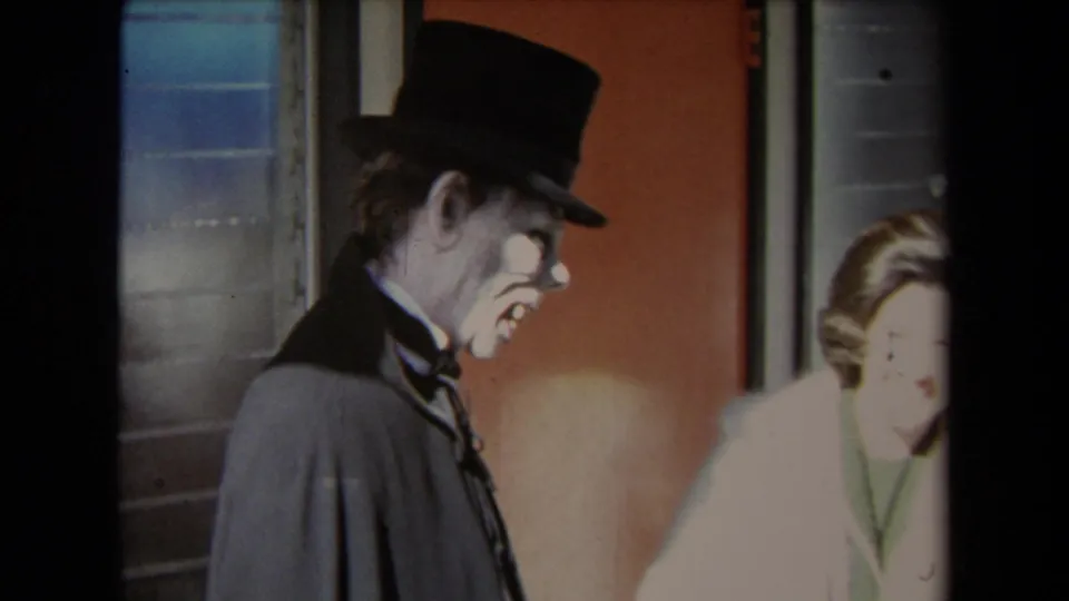 1967 Los Angeles home-movie frame showing a grotesque white-faced costumed man in a top hat beside a woman near a window and orange wall.