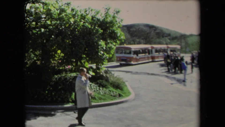 Outdoor 1967 Los Angeles 8mm frame showing a man in a light coat near leafy landscaping, pavement, hills, and a red-and-white tour train.