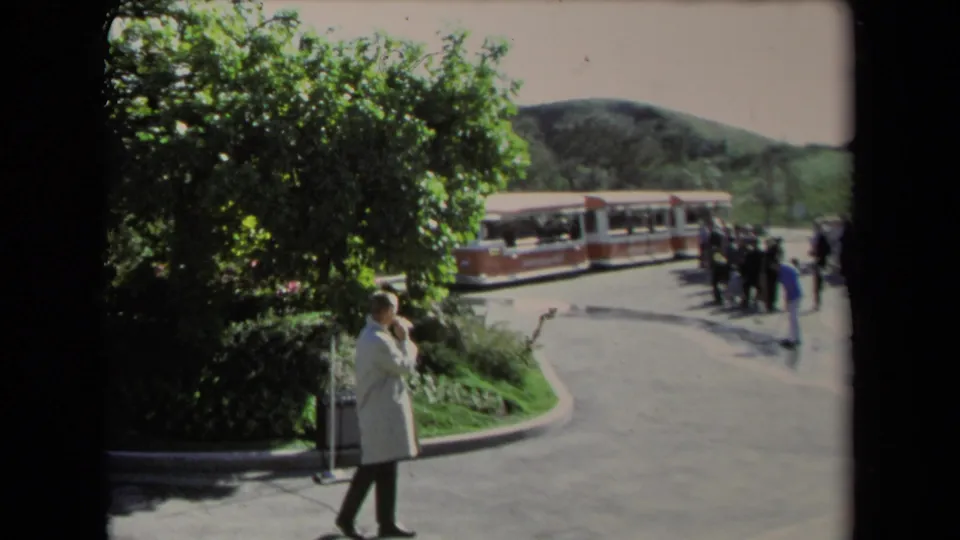1967 Los Angeles home-movie frame of a man by landscaped greenery, with a red-and-white tour train and small group of visitors beyond him.