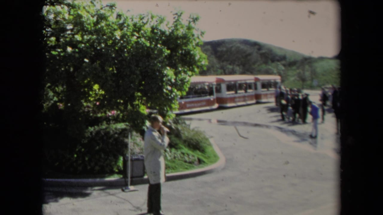 Man in a light coat speaking at a microphone near a red-and-white tour train and trees.