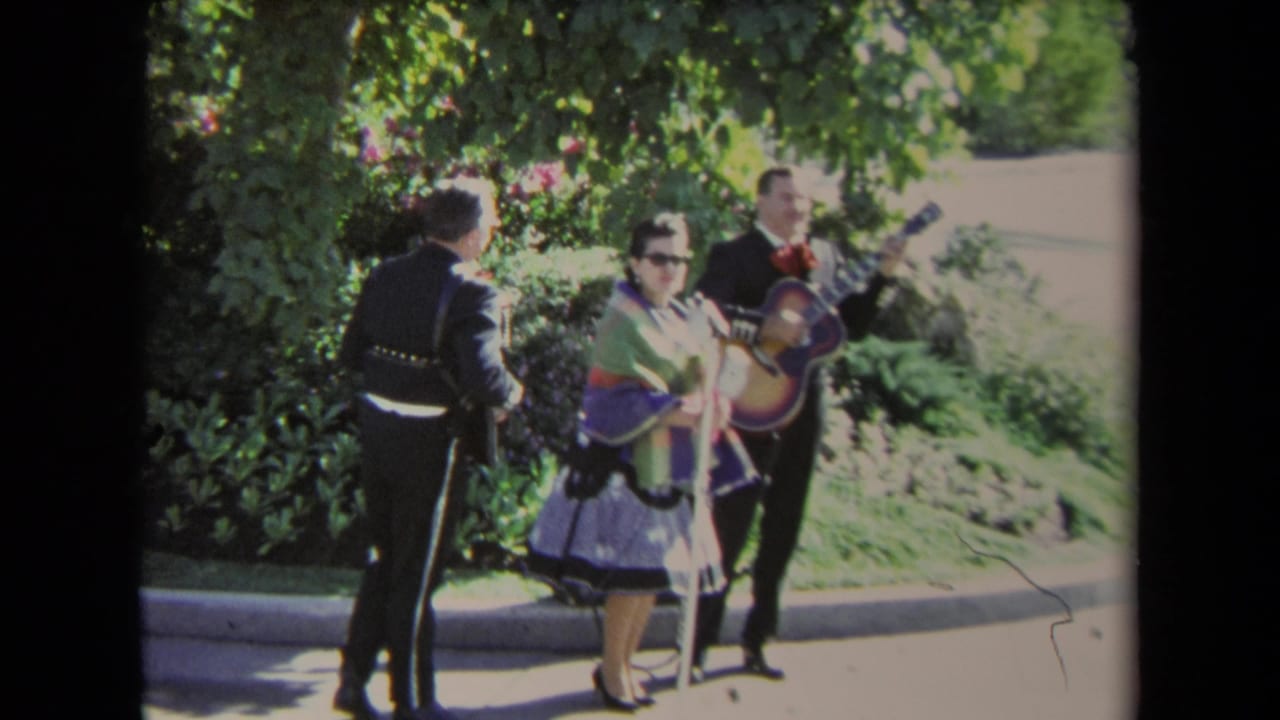 1967 Los Angeles frame of three mariachi-style performers outdoors, including a woman in a colorful dress and two men in dark suits.