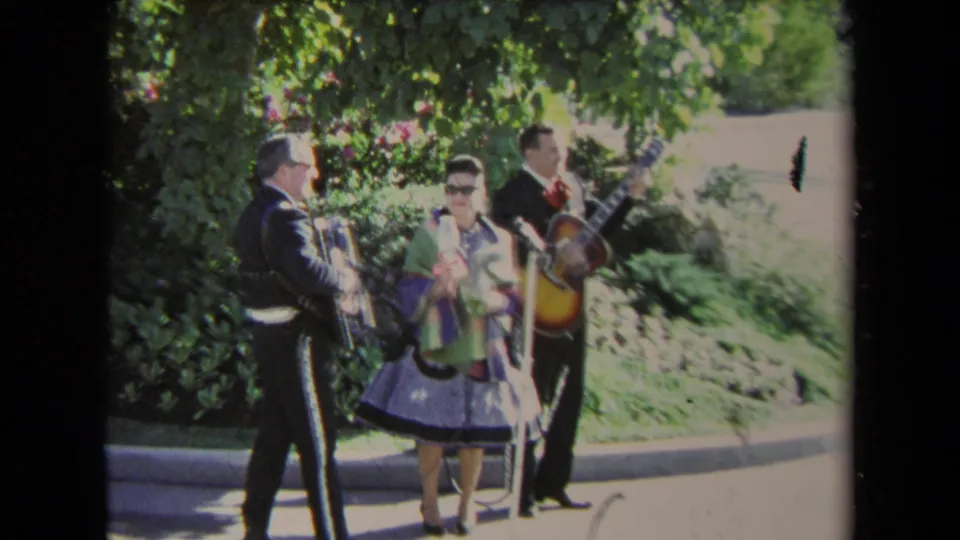 1967 Los Angeles 8mm frame showing a mariachi-style trio on a sidewalk, with accordion and guitar visible against trees and shrubs.