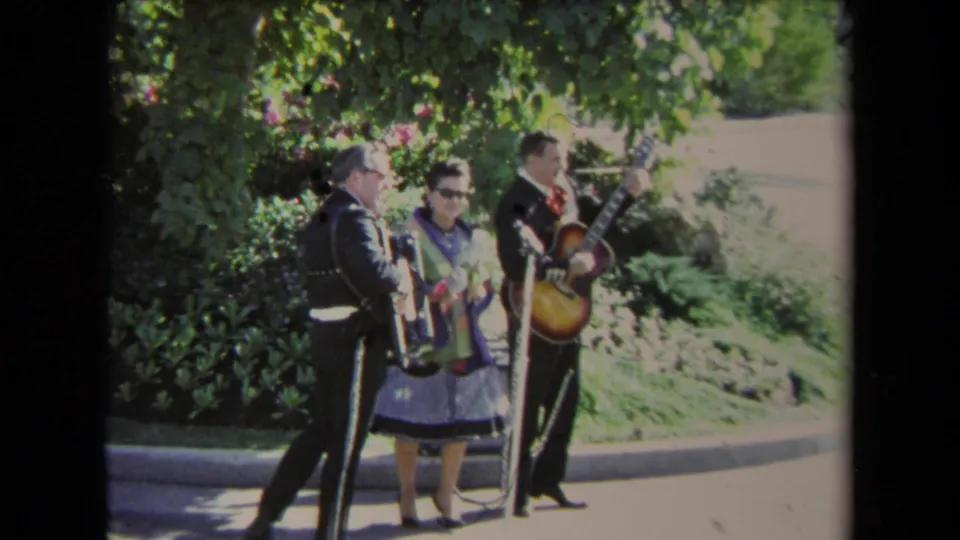 1967 Los Angeles frame of the mariachi-style trio gathered near microphones, with a guitarist, accordion player, and woman performer outdoors.