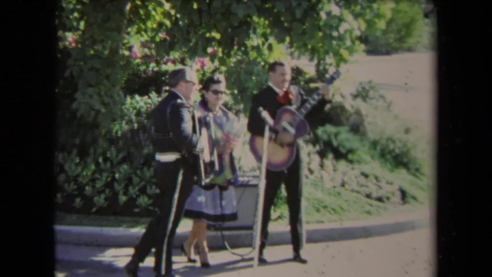1967 Los Angeles home-movie frame showing mariachi musicians with guitar and accordion under trees as a woman watches nearby.