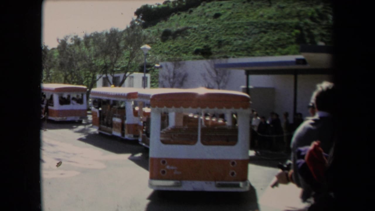 1967 Los Angeles frame showing orange and white tourist trolleys parked near a hillside station area with visitors nearby.