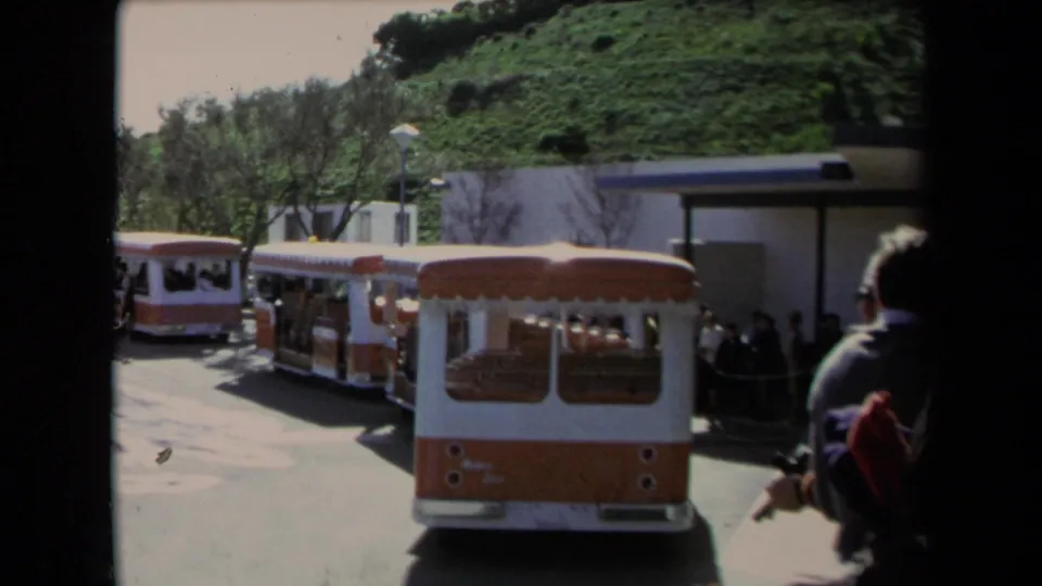 1967 Los Angeles frame showing orange and white tourist trolleys parked near a hillside station area with visitors nearby.