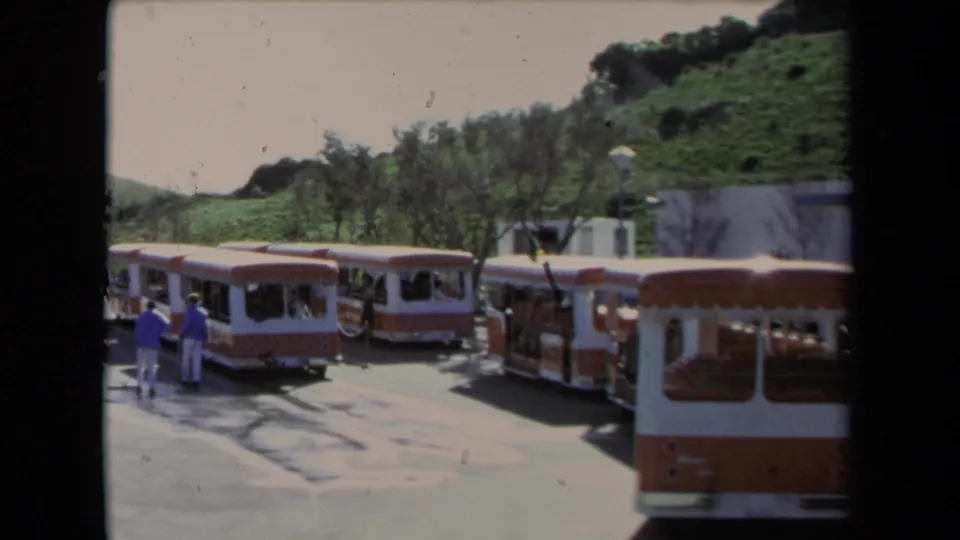 1967 Los Angeles home-movie frame with parked tourist trolleys, visitors walking nearby, trees, and a hillside behind the station.
