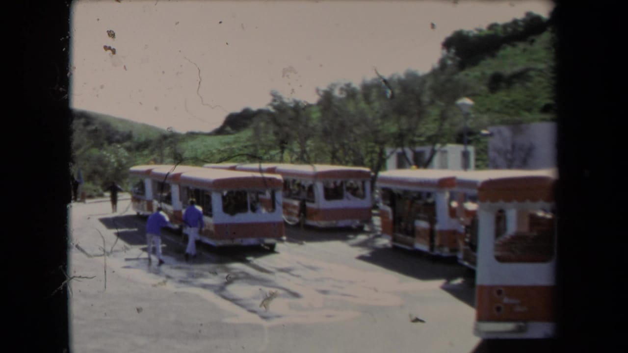 1967 Los Angeles home-movie frame showing orange-and-white open-air tour buses parked near visitors on a sunny paved lot.