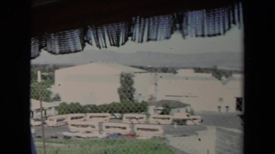 1967 Los Angeles home-movie frame looking from inside a vehicle past a curtain toward buses, fencing, and low industrial buildings.