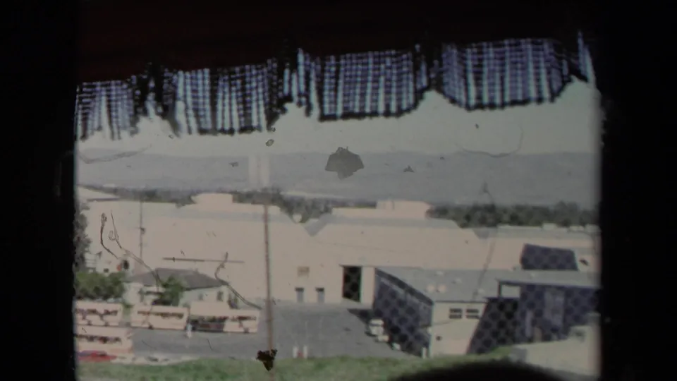 1967 Los Angeles frame from inside a vehicle, with a curtain crossing the view of fencing, low buildings, and parked vehicles.