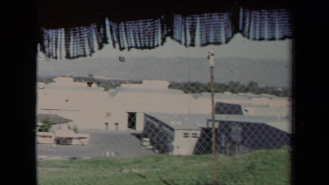 1967 Los Angeles 8mm frame looking past a checkered curtain toward fencing, low buildings, grass, and hazy hills.