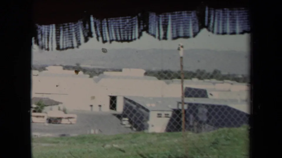1967 Los Angeles 8mm frame looking past a checkered curtain toward fencing, low buildings, grass, and hazy hills.