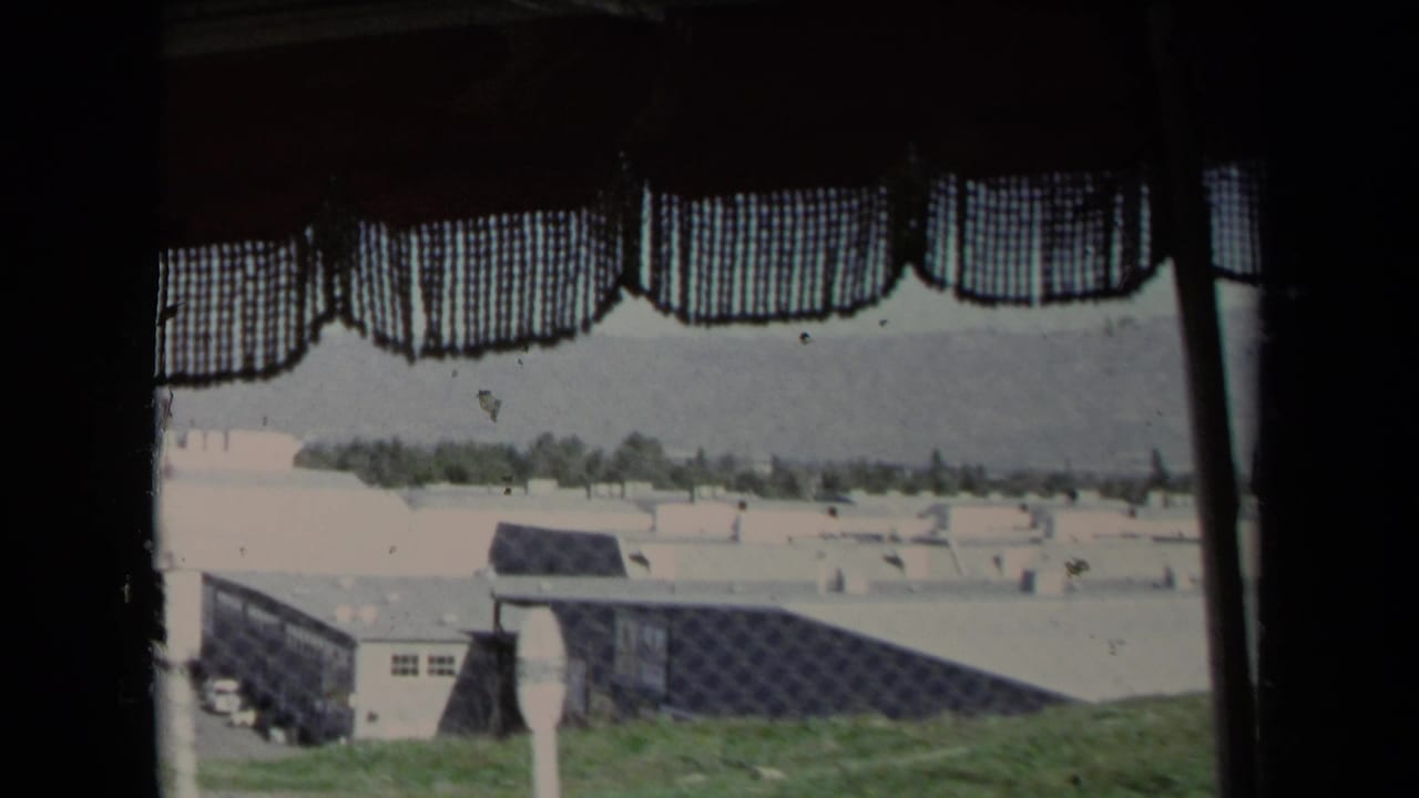 A faded 1967 Los Angeles home-movie frame shows fenced low buildings and distant trees through a curtained vehicle window.