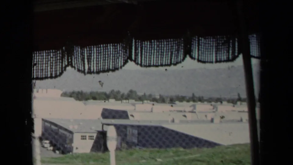 A faded 1967 Los Angeles home-movie frame shows fenced low buildings and distant trees through a curtained vehicle window.