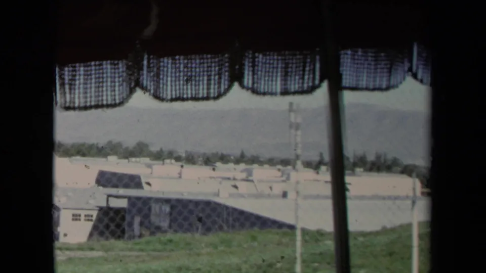 Grainy 8mm view in Los Angeles, 1967, looking through a curtained window at fenced low buildings and a grassy foreground.