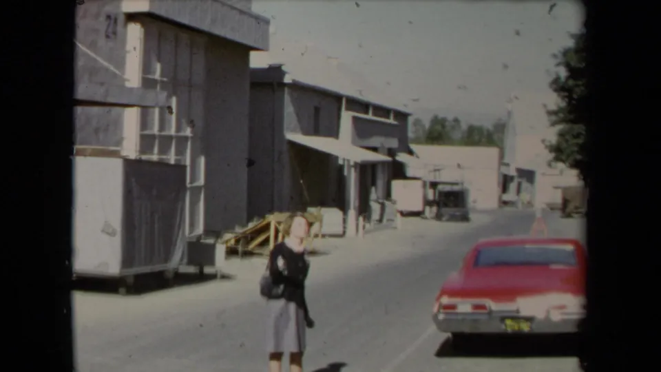 1967 Los Angeles street frame with a woman walking near warehouse-style buildings and a red car parked along the road.