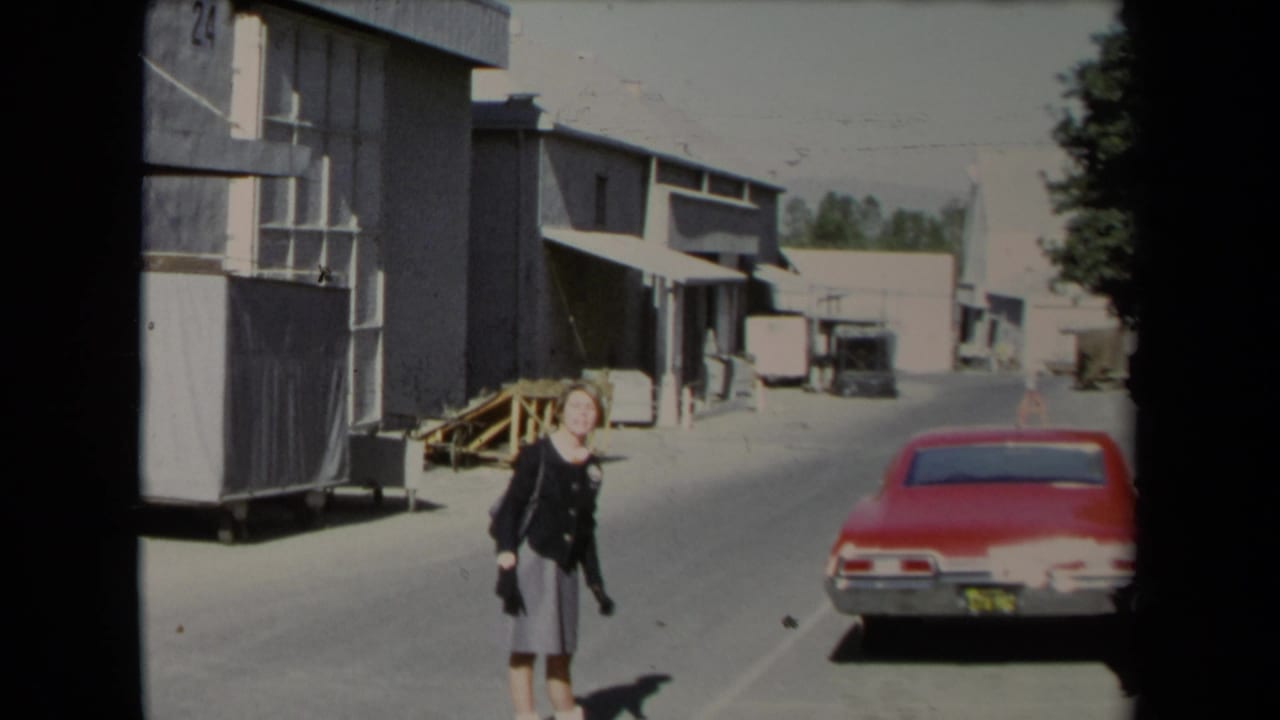 In 1967 Los Angeles footage, a woman walks through an industrial alley beside warehouse buildings and a red car.
