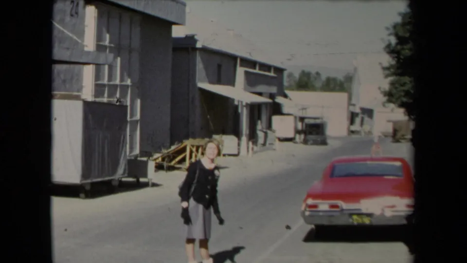 In 1967 Los Angeles footage, a woman walks through an industrial alley beside warehouse buildings and a red car.