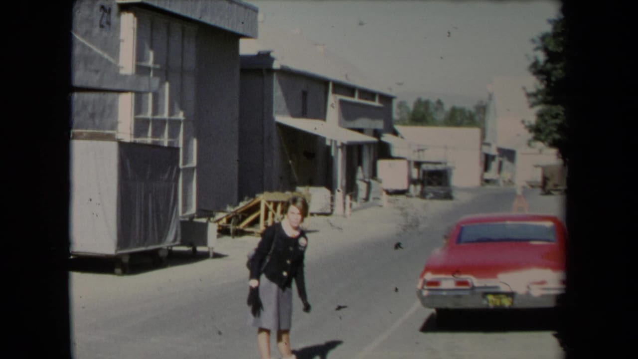 The woman moves along a paved industrial area in Los Angeles, with a red car and warehouse structures visible behind her.