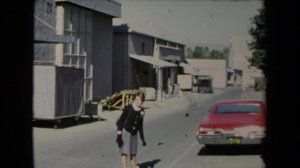 The woman moves along a paved industrial area in Los Angeles, with a red car and warehouse structures visible behind her.