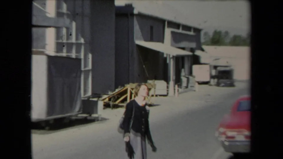 A tense-looking woman gestures as she walks through a Los Angeles industrial alley with warehouse facades and a red car in view.