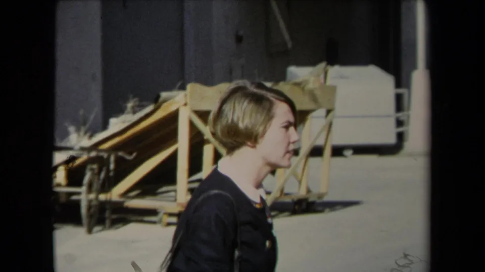 A closer side view shows the woman in a dark jacket and gray skirt passing a loading ramp in a Los Angeles industrial area.