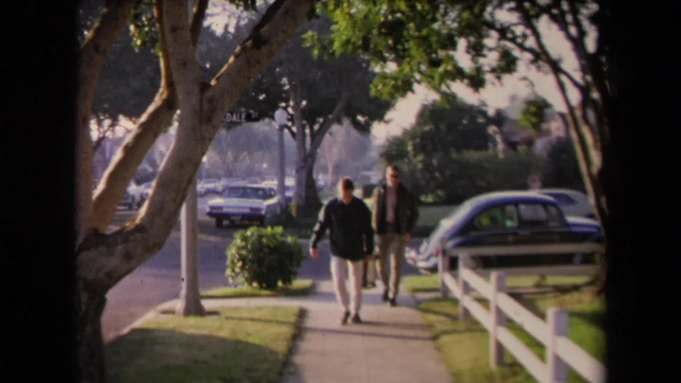 In Los Angeles home-movie footage, two men walk along a sunny sidewalk with trees, parked mid-century cars, grass, and a white rail fence nearby.