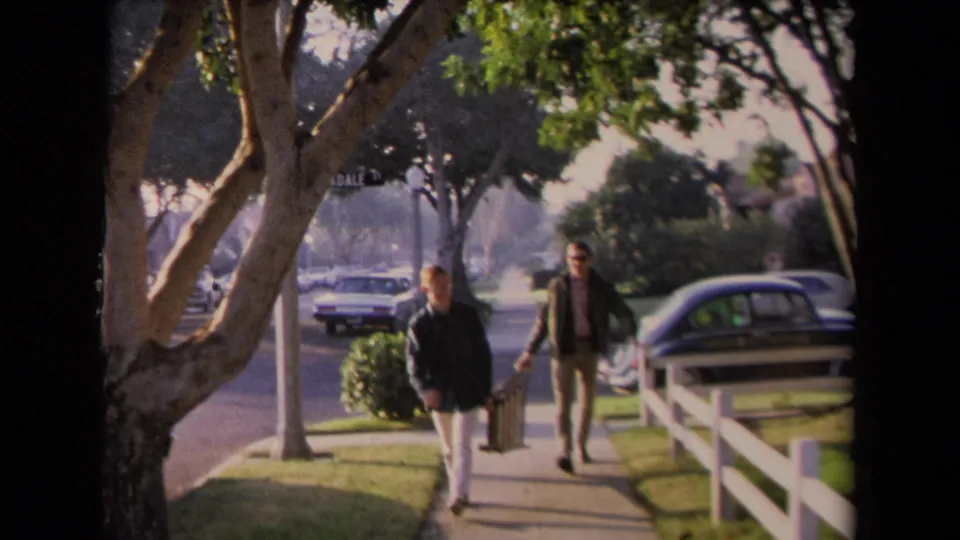 Two men carry a ladder together along a 1967 Los Angeles sidewalk, with trees, parked cars, grass, and a white fence in the background.