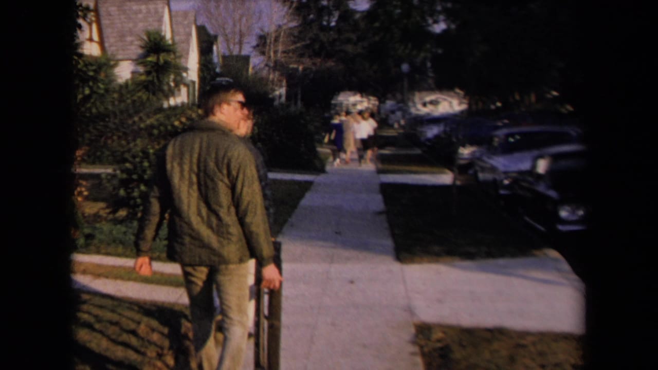 Man in green jacket walks on a suburban sidewalk carrying a long object.