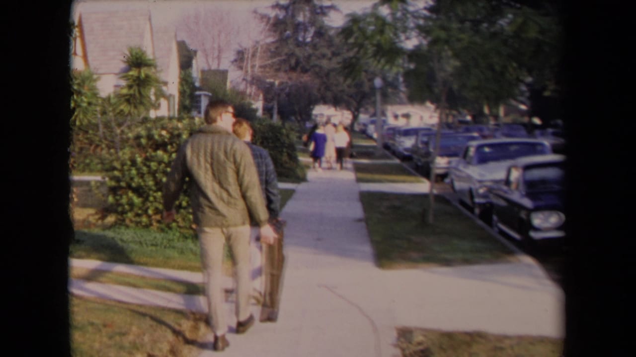 Two men walk down a Los Angeles suburban sidewalk in 1967, with lawns, hedges, trees, and mid-century cars along the street.