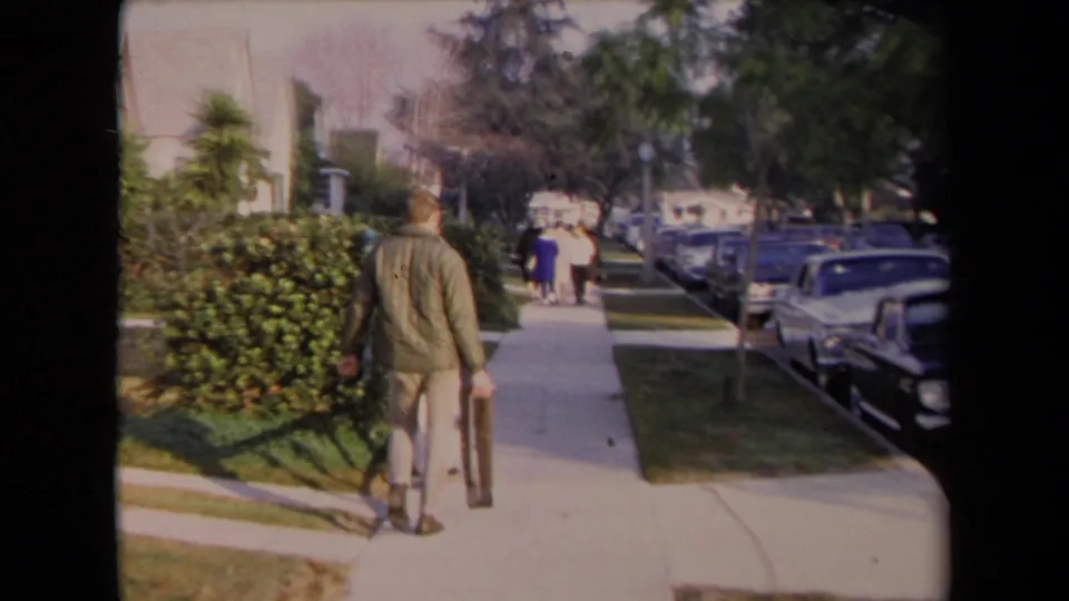 A man seen from behind walks along a 1967 Los Angeles residential sidewalk, carrying a long object past hedges and parked cars.