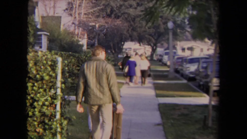 A 1967 Los Angeles sidewalk view shows a man walking away toward other pedestrians, with lawns, hedges, and parked cars visible.