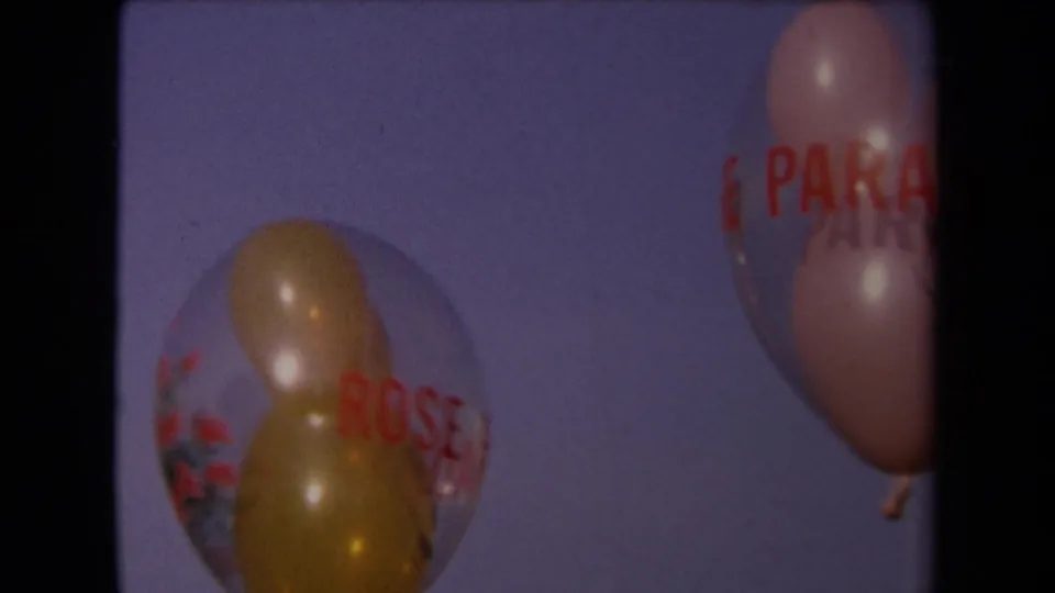 A clear balloon with red lettering and a pink balloon move through the soft blue sky in the home-movie frame.