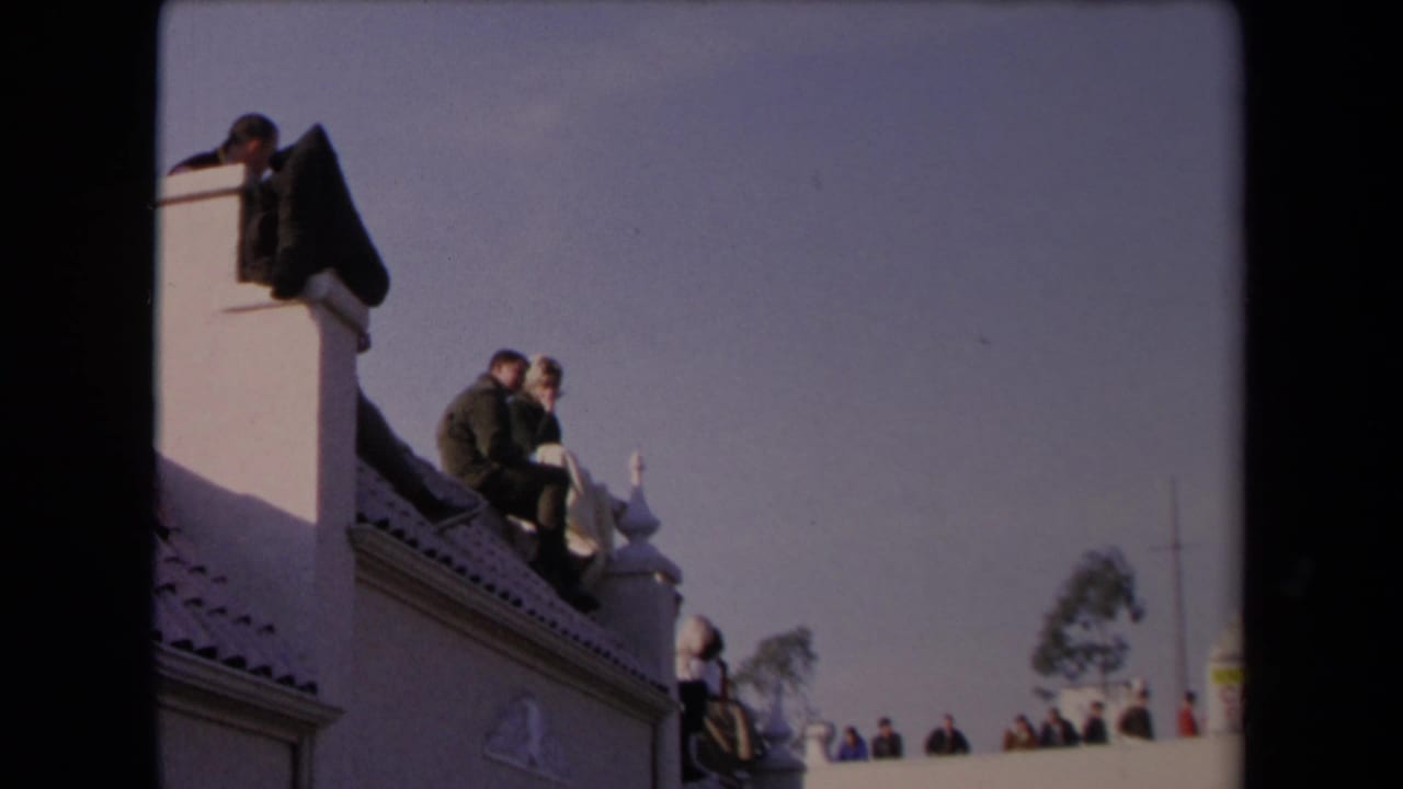 1967 Los Angeles 8mm frame showing a couple on a white rooftop ledge, with distant spectators under a pale sky.