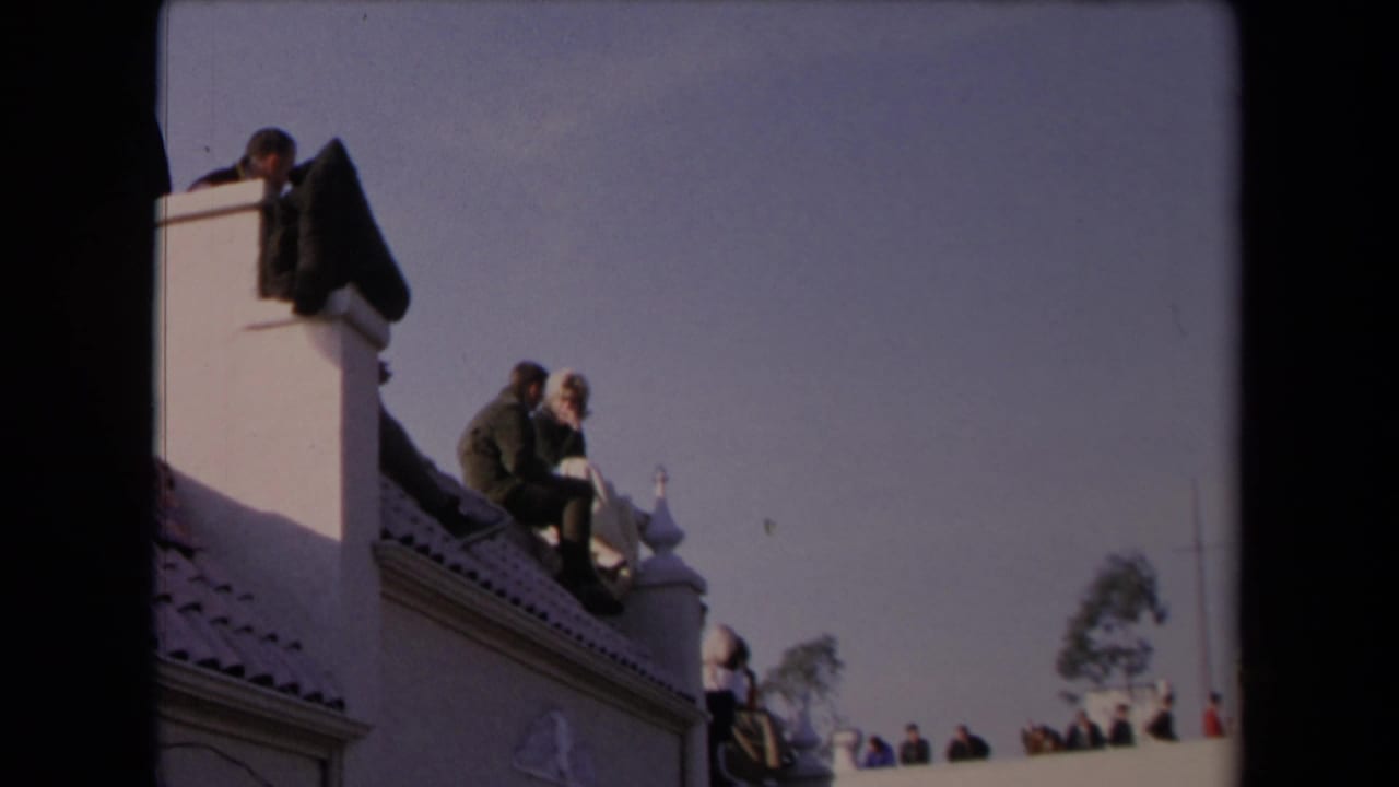 1967 Los Angeles rooftop scene with a seated couple, white stucco architecture, red tile roof, and spectators farther back.
