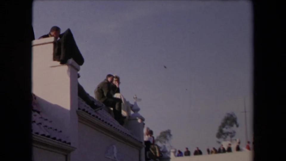 1967 Los Angeles home-movie frame looking up at a couple on a tiled rooftop, with spectators and soft film grain visible.
