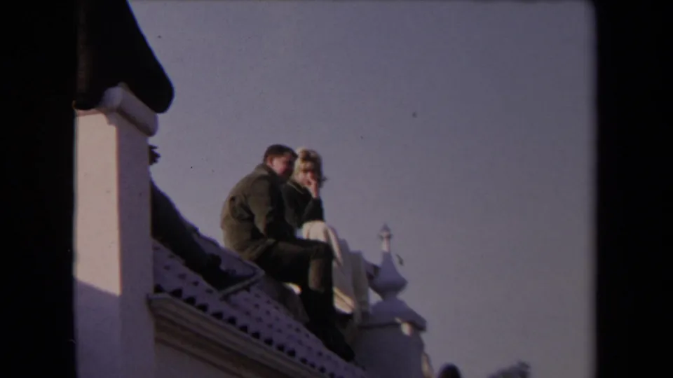 Closer 1967 Los Angeles 8mm frame of a man in a dark jacket and a blonde woman seated on a rooftop ledge.