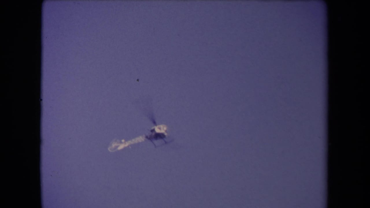 Small helicopter flying against a blue sky within a dark 8mm film frame.