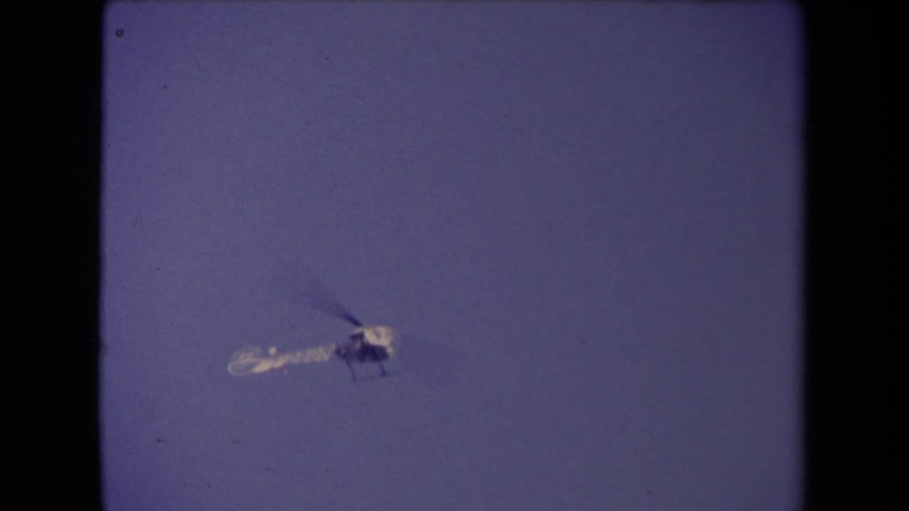 The helicopter continues across the clear sky, framed by the soft grain and borders of 8mm footage.