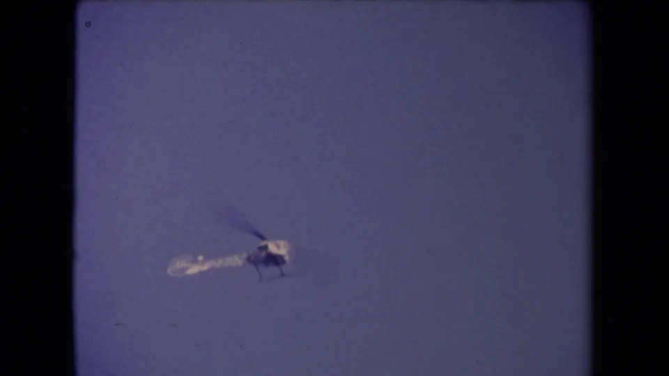 The helicopter continues across the clear sky, framed by the soft grain and borders of 8mm footage.