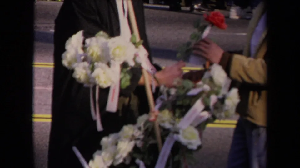 In Los Angeles, adults in formal clothing handle white floral wreaths and a red rose beside road markings.