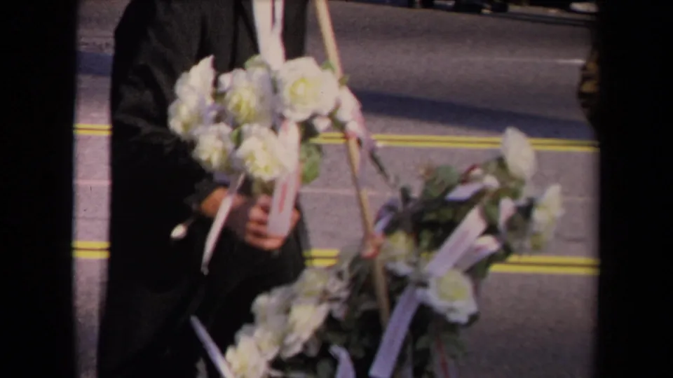 A person in dark formal clothing carries white floral wreaths with ribbons on a Los Angeles street.