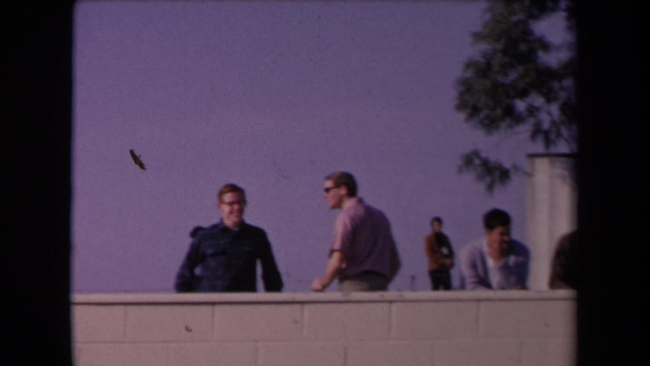 1967 home-movie frame of men behind a rooftop or raised wall in Los Angeles, including a dark-uniformed officer and casual spectators.