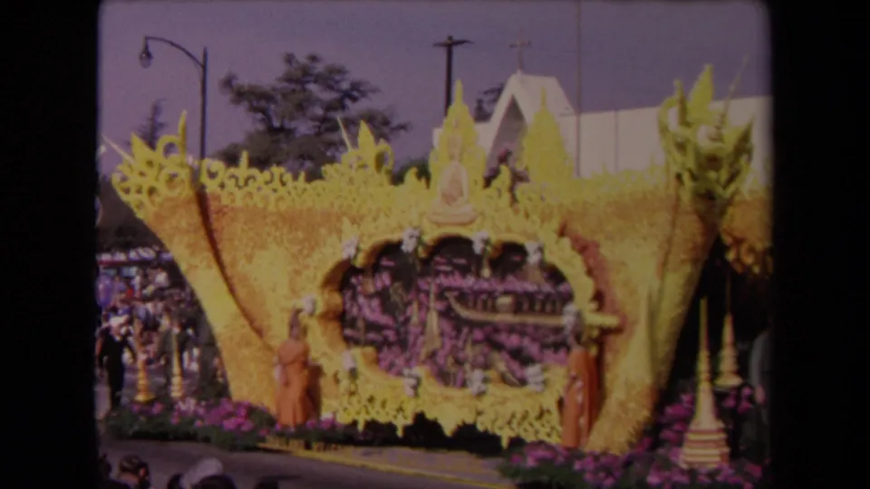 Home-movie frame from 1967 Los Angeles showing the yellow floral float, Buddha figure, spectators, and a church steeple beyond the route.