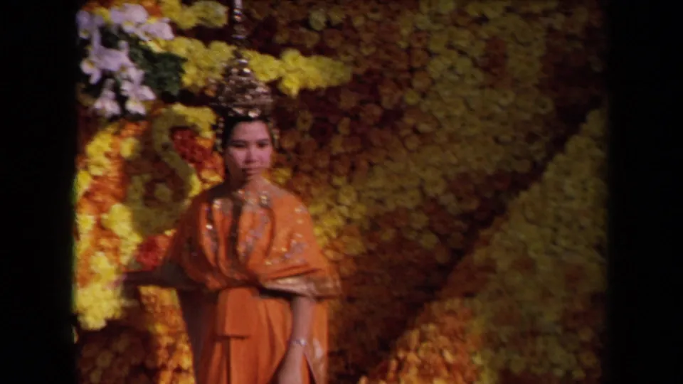 1967 Los Angeles parade frame showing a woman in orange traditional dress standing on or near the floral float backdrop.
