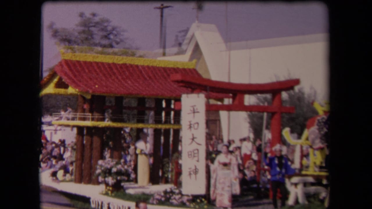 1967 Los Angeles parade float seen from the side, with the pavilion roof prominent, a vertical character sign, red torii gate, flowers, and visible participants.