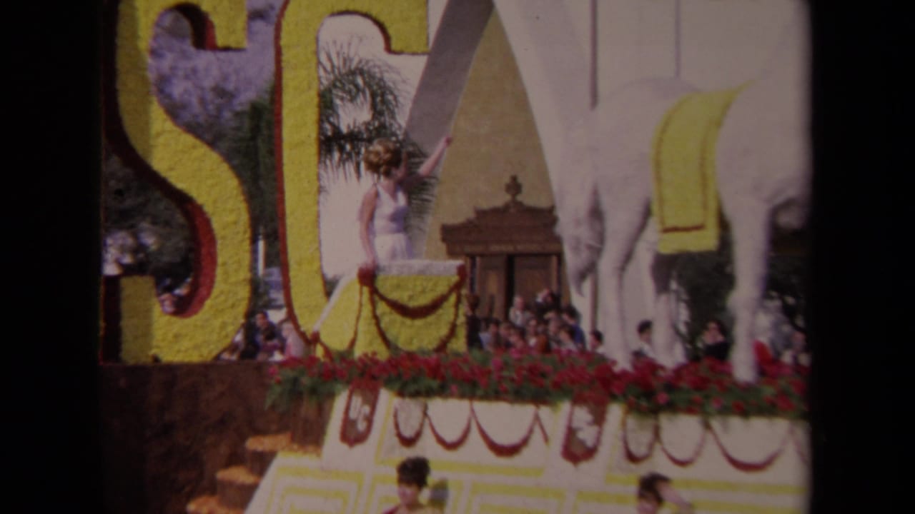 1967 Los Angeles parade frame showing a woman in white on a floral float with large yellow lettering, a white horse figure, and spectators below.