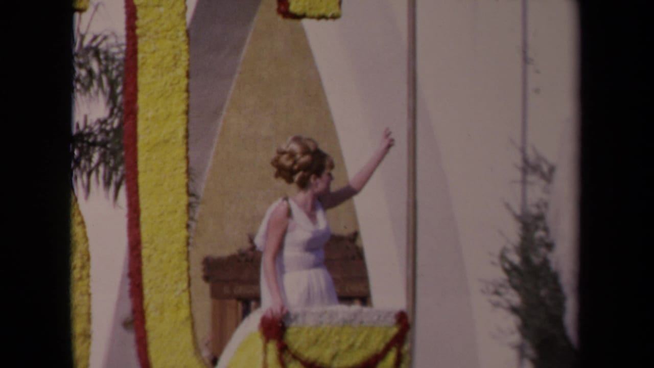 1967 Los Angeles parade view of a woman waving from a decorated float, with yellow display lettering, floral trim, and a white horse figure.