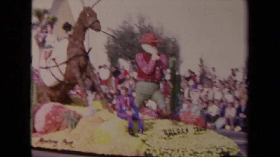1967 Los Angeles parade frame showing a cowboy-themed floral float with a horse figure, red-hatted figure, seated man, and spectators.