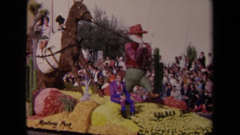 1967 Los Angeles parade view of a floral cowboy-themed float, horse figure, seated man in purple, and packed spectators along the route.